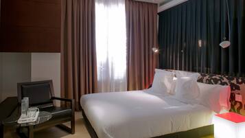 A hotel room with a bed, a chair, a table with glasses and a bottle, and a nightstand with a lamp.