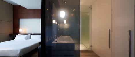 Una habitación de hotel moderna con una cama grande, un baño con paredes de vidrio y un diseño minimalista.