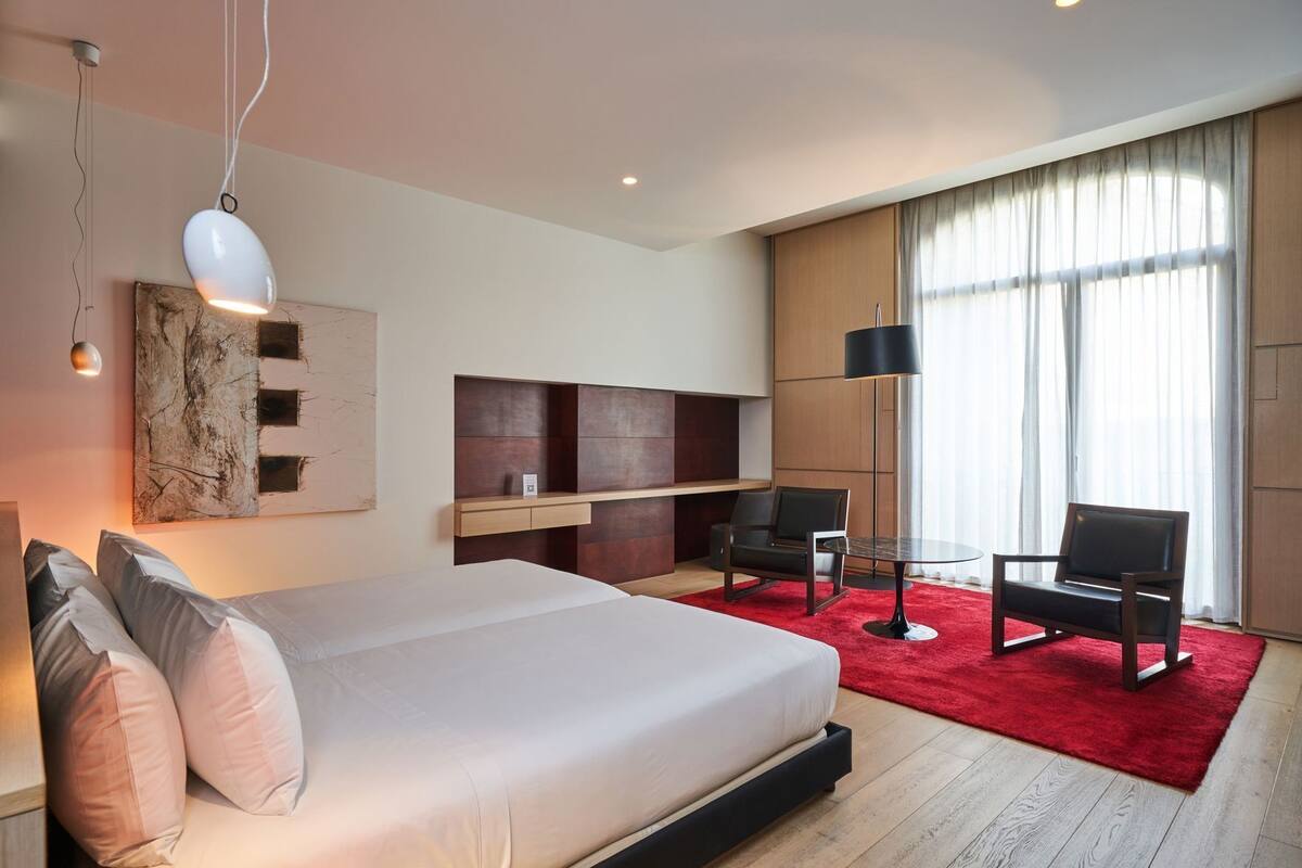 Ein modernes Hotelzimmer mit einem großen Bett, einem Schreibtisch, zwei Stühlen, einem roten Teppich und einem großen Fenster mit Vorhängen.
