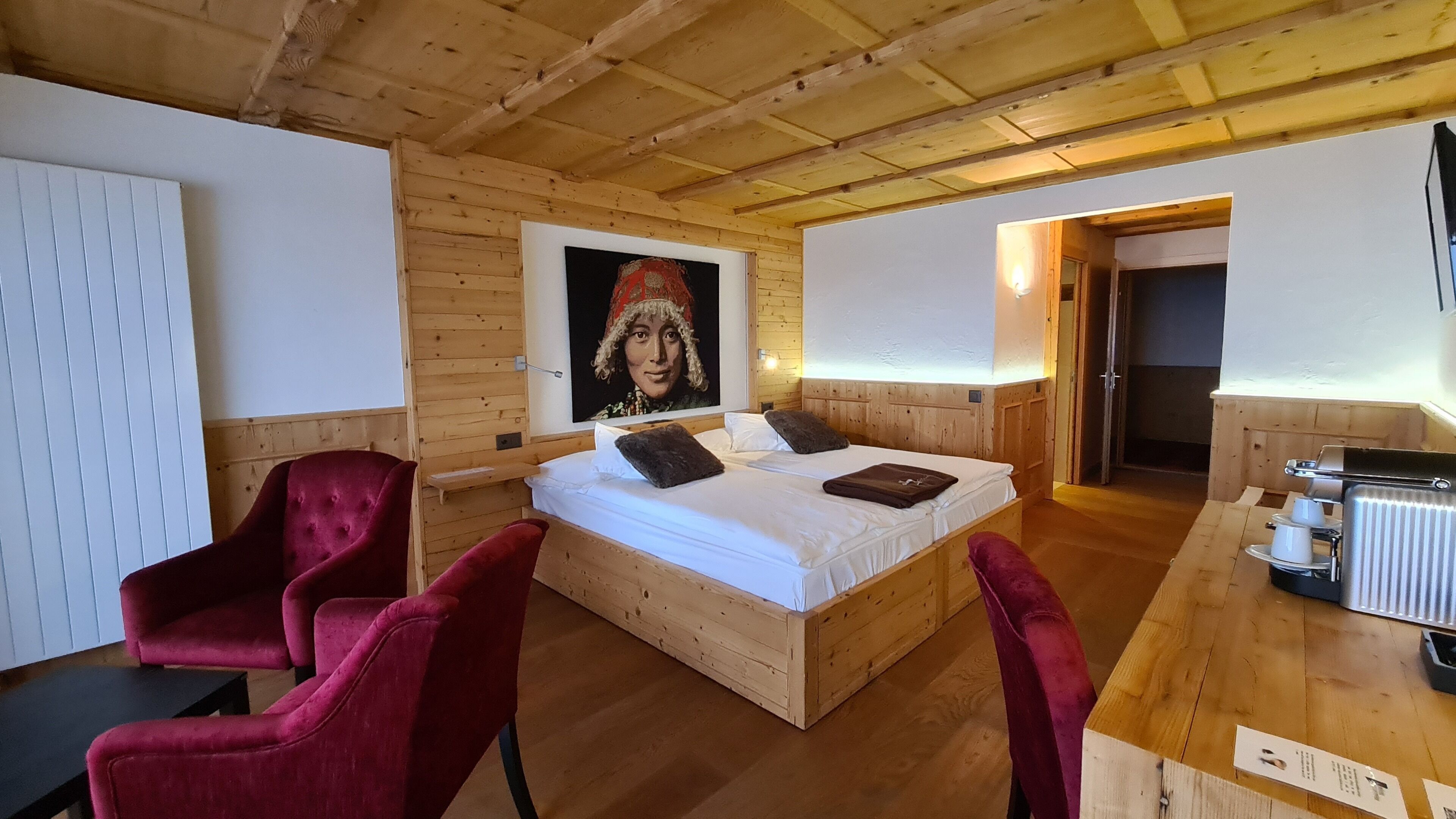 A wooden-paneled room with a bed, two red chairs, a table with a kettle, and a painting on the wall.