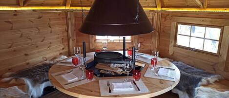 Interior de una cabaña de madera con una mesa redonda, una lámpara colgante y bancos cubiertos de piel.