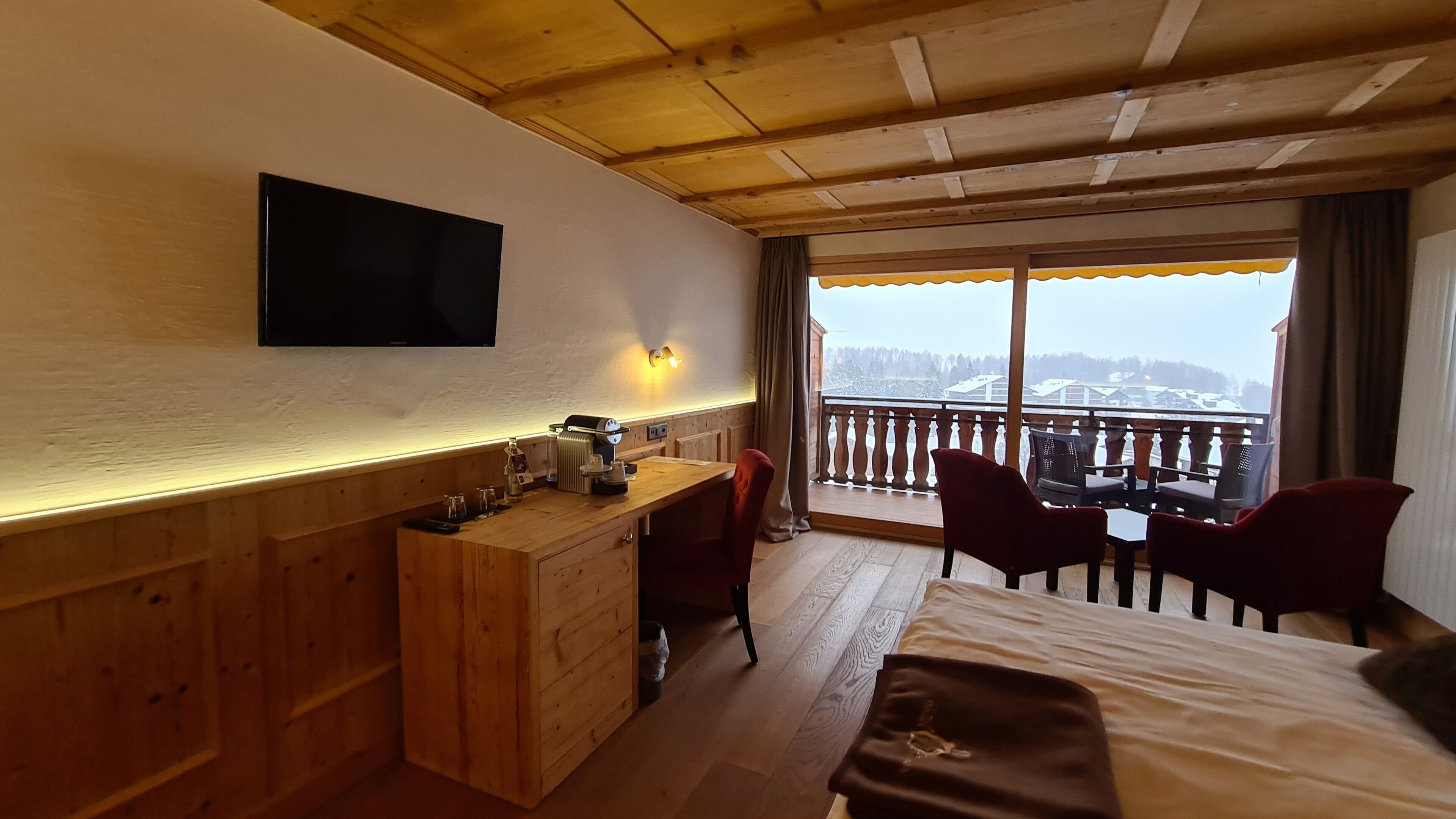 A hotel room with a wooden desk, a TV mounted on the wall, a balcony with a view of the snowy landscape, and a bed with a brown blanket.