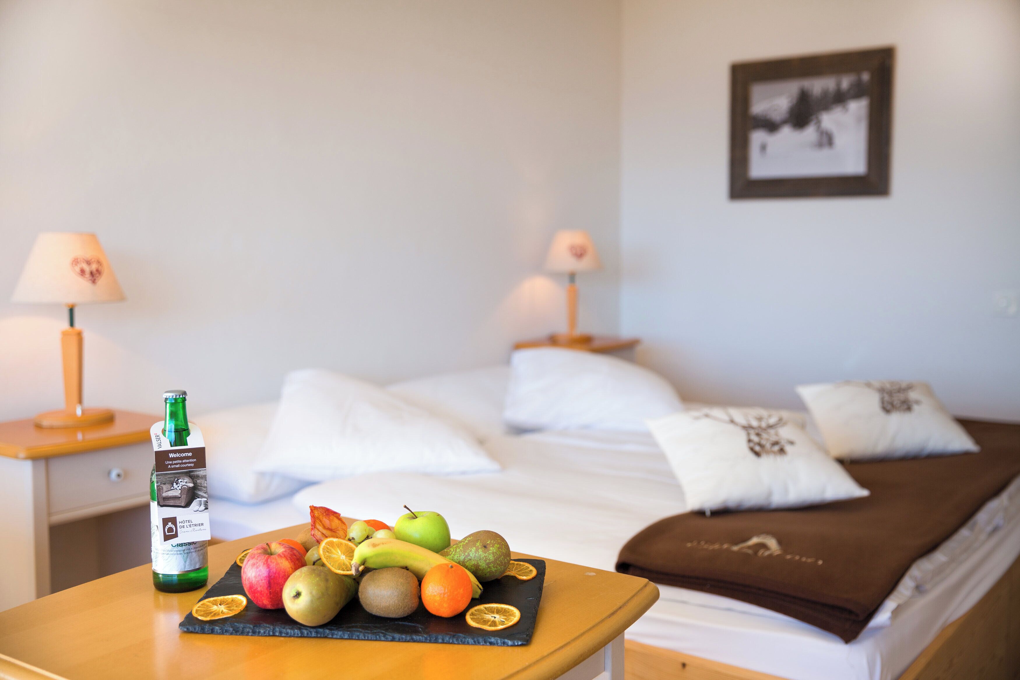 Habitación de hotel con cama, almohadas, mesita de noche con una botella y fruta, y un cuadro colgado en la pared.
