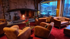 A cozy lounge area with leather sofas, a fireplace, and a view of a mountain resort. - Hôtel de l'Etrier (Crans-Montana)