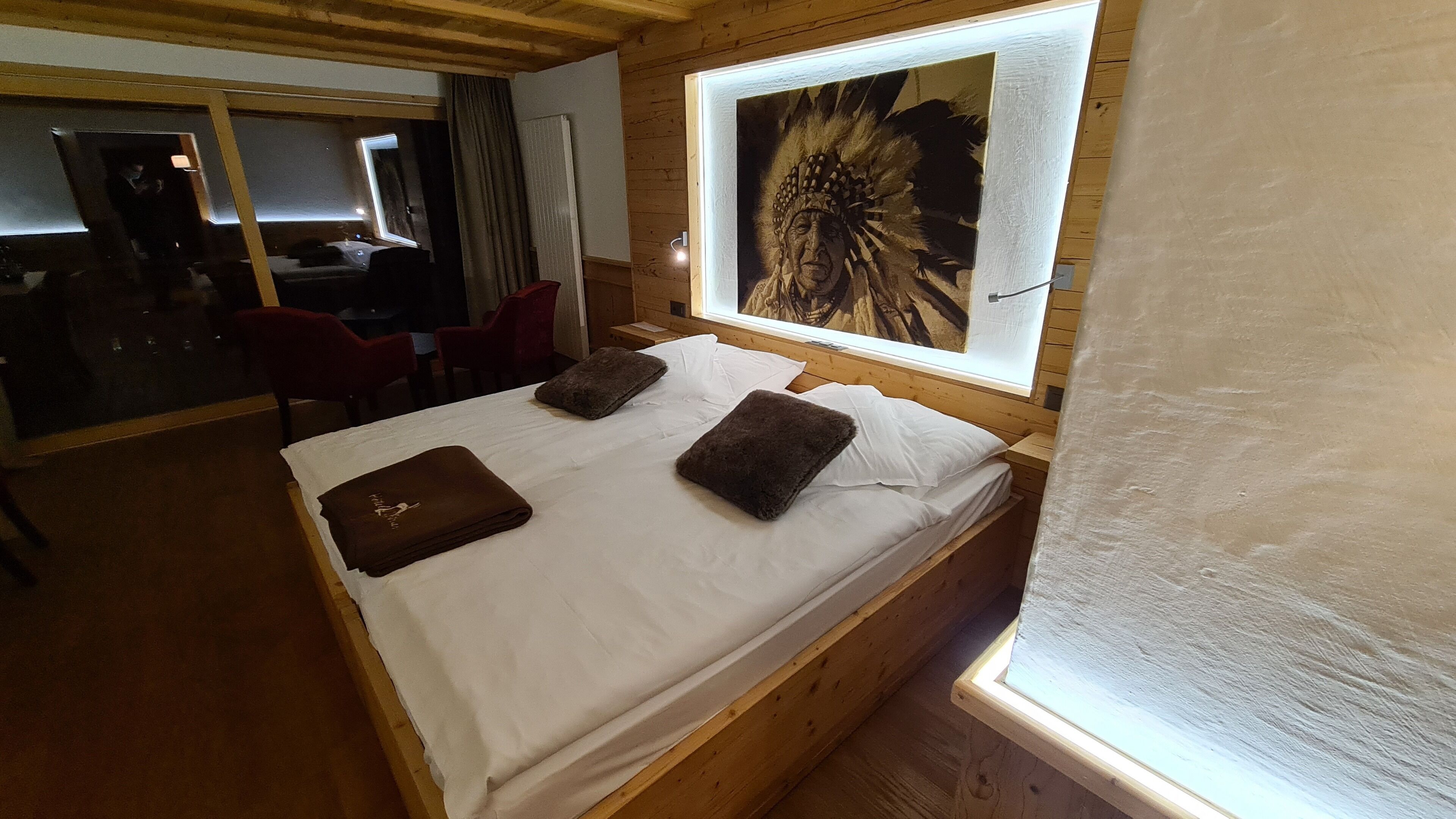 A wooden bed with white linens and two brown pillows, a painting above the bed, and a mirror on the wall.