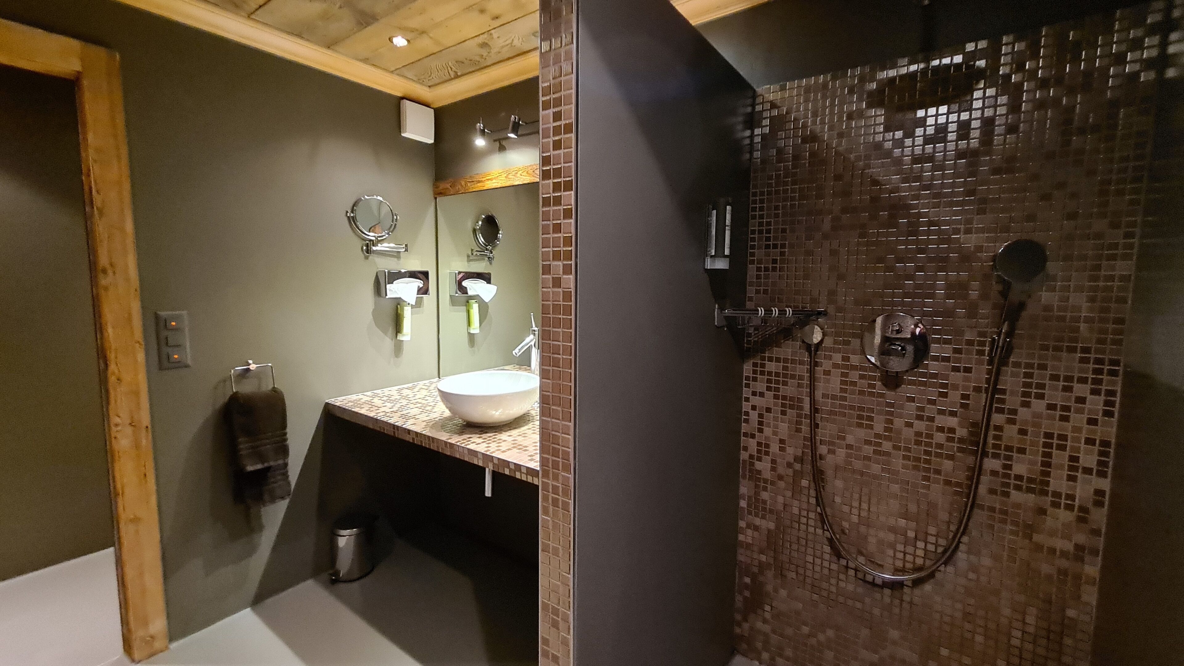 A bathroom with a wooden ceiling, a sink with a mirror, and a shower with mosaic tiles.