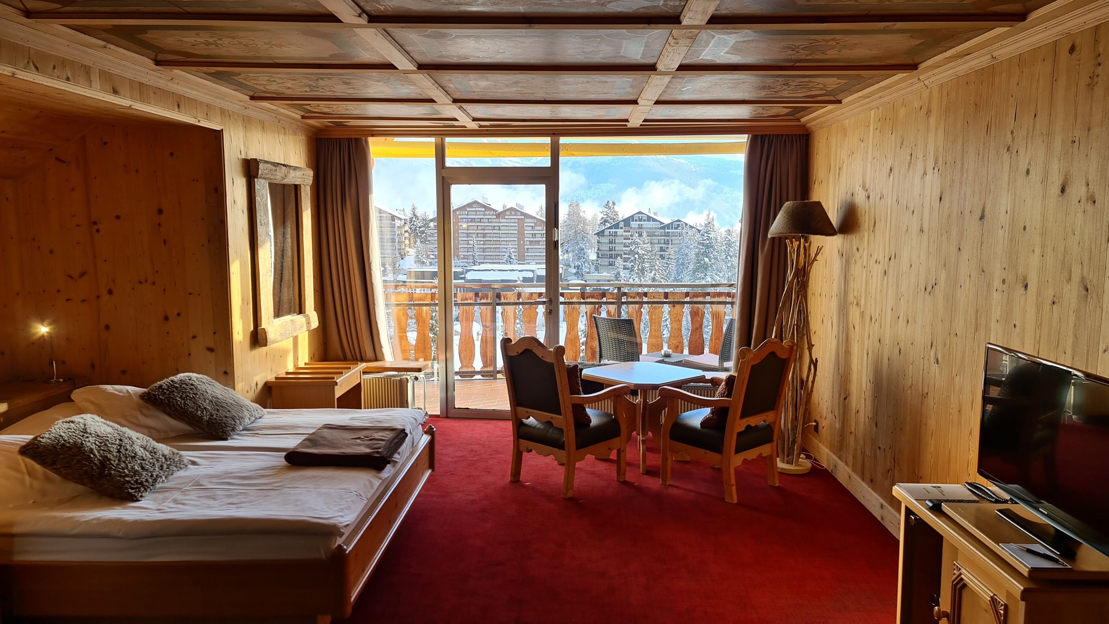 A hotel room with a bed, a desk with a chair, a TV, and a balcony with a view of buildings and snow.