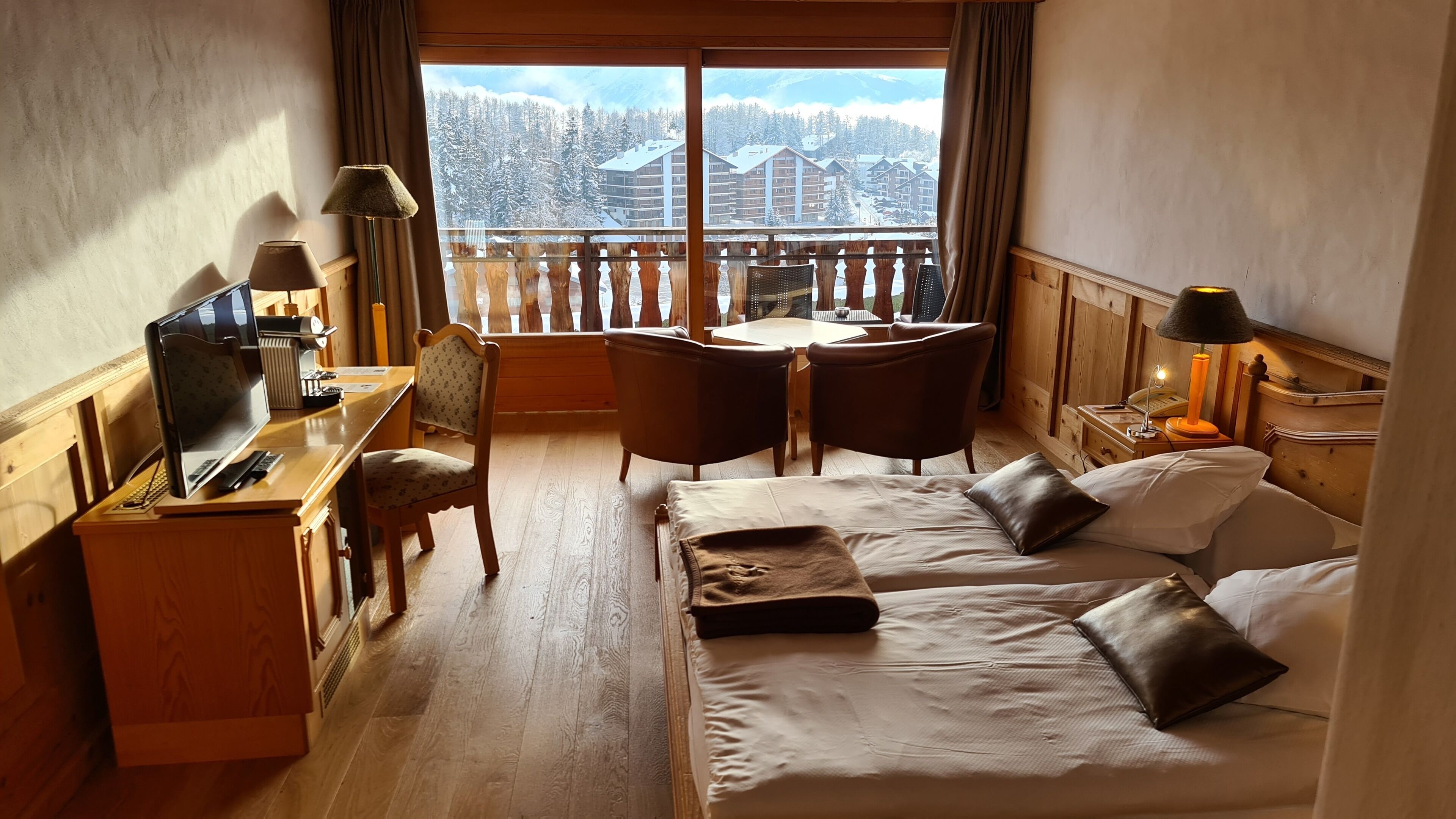 A hotel room with a bed, desk, chair, and a view of snow-covered mountains.