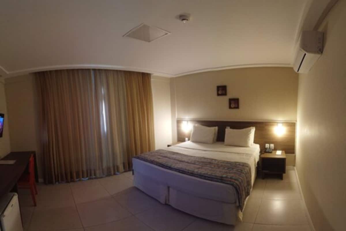 Habitación de hotel con una cama grande, un escritorio con computadora, una mesita de noche con lámpara y una ventana con cortinas.