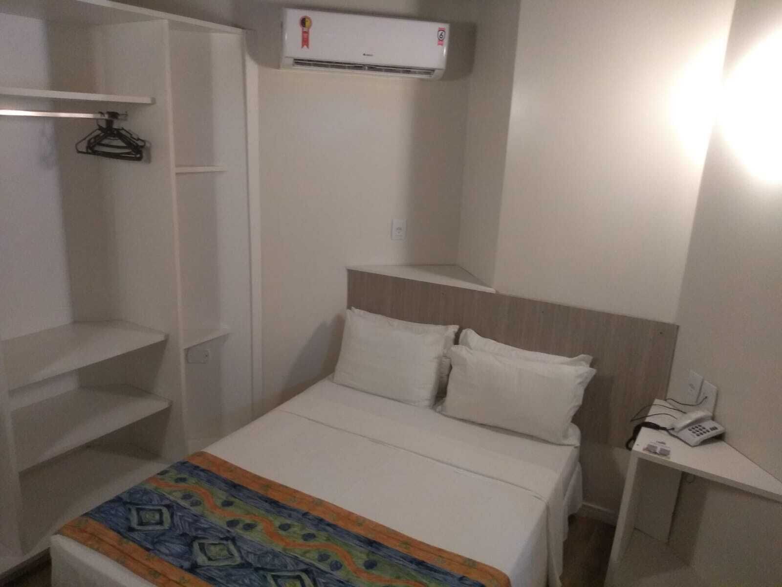 A hotel room with a bed, a bedside table with a phone, a wardrobe, and an air conditioning unit.
