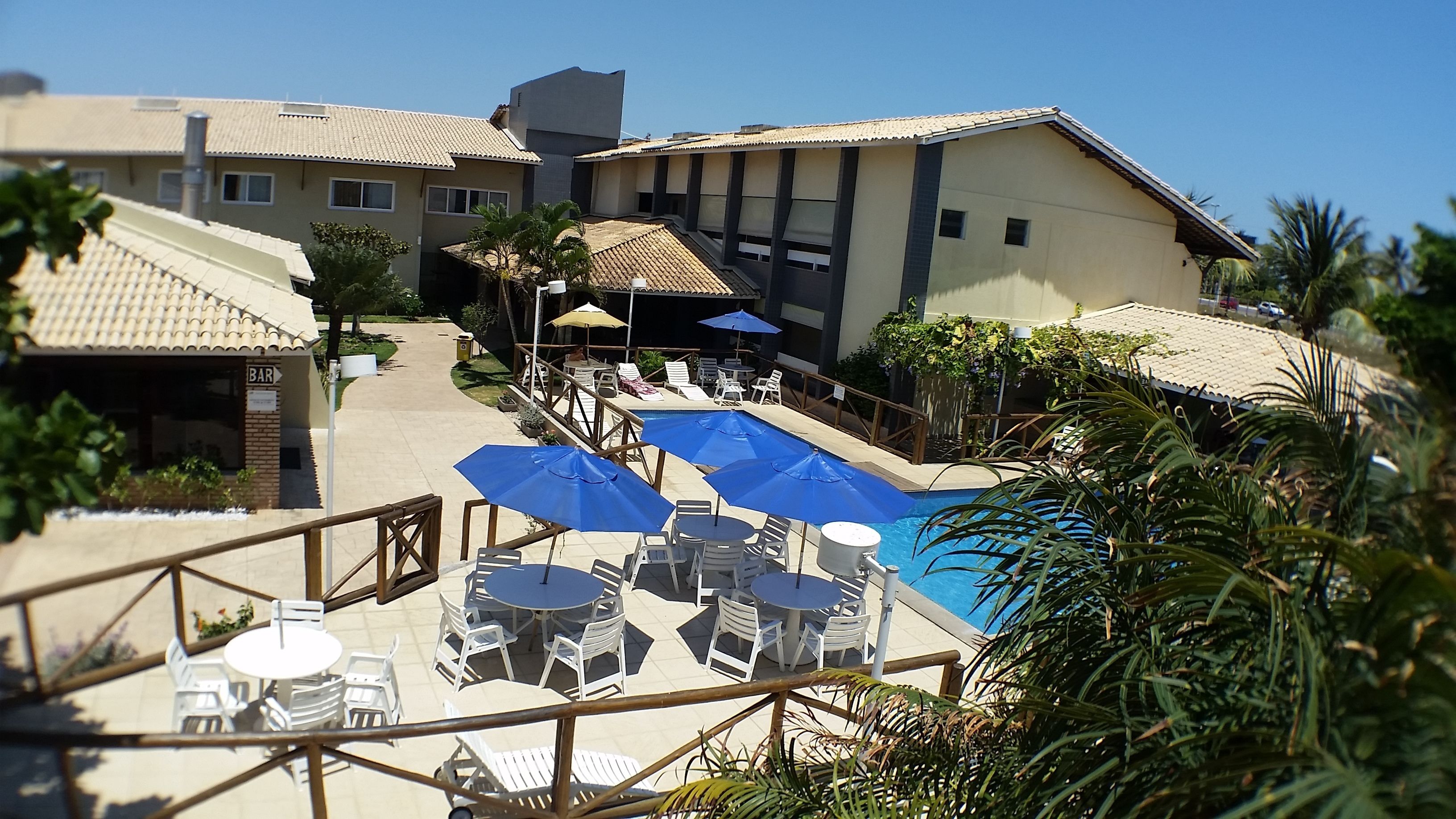Un área junto a la piscina con varias mesas y sombrillas azules, rodeada de edificios con tejados de tejas y una zona de bar.