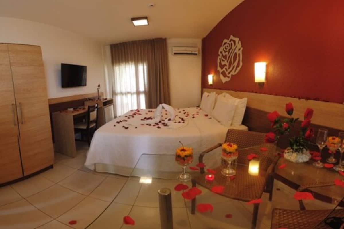 Habitación de hotel con cama, escritorio, televisión y zona de comedor con pétalos de rosa y flores.