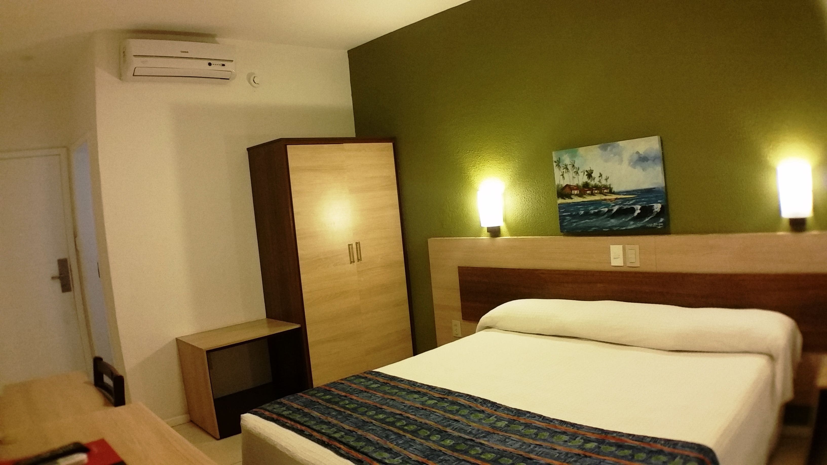 A hotel room with a bed, a wardrobe, a small table, and a painting on the wall.