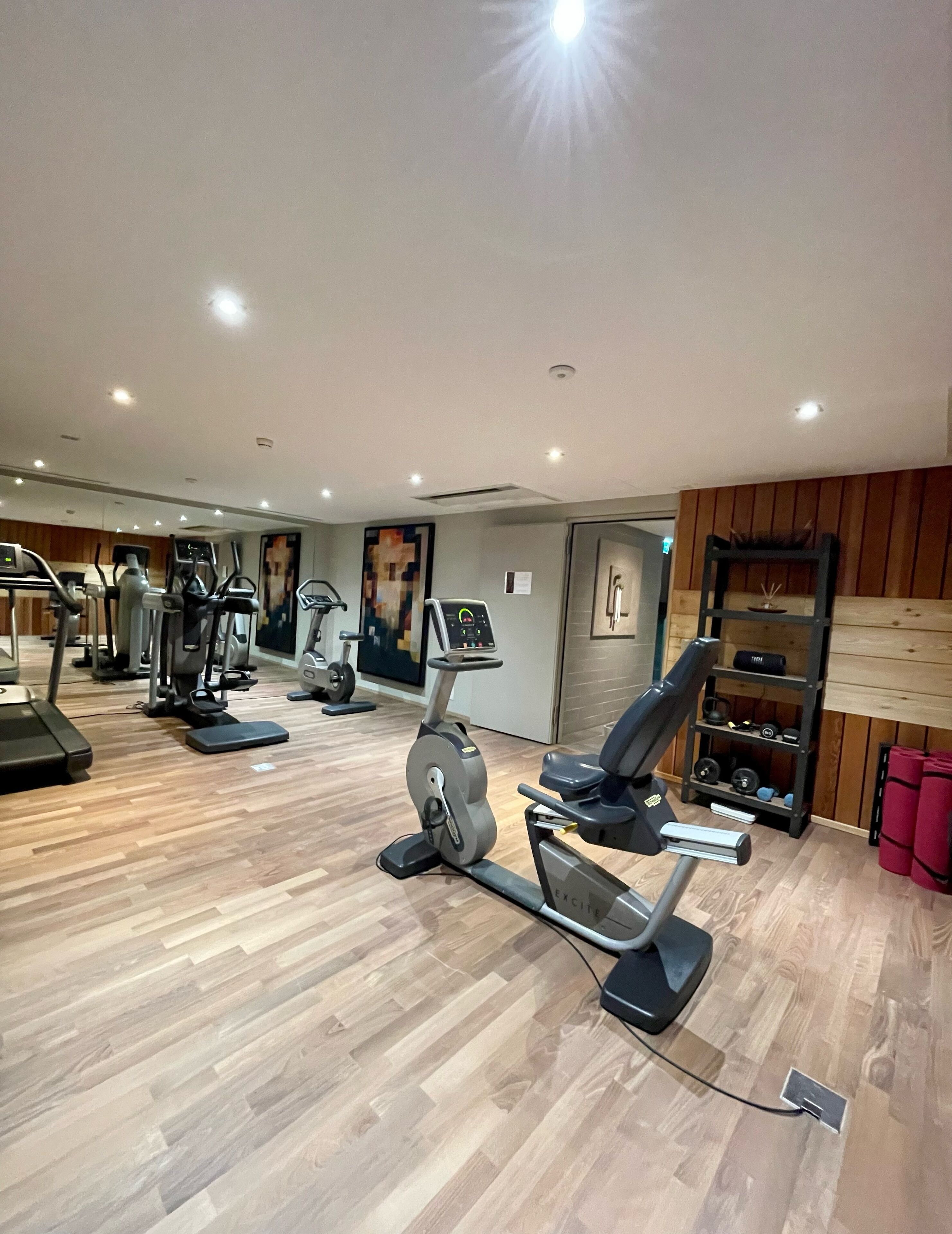 Ein Fitnessraum mit unterschiedlichen Trainingsgeräten, Holzboden und einer Spiegelwand.