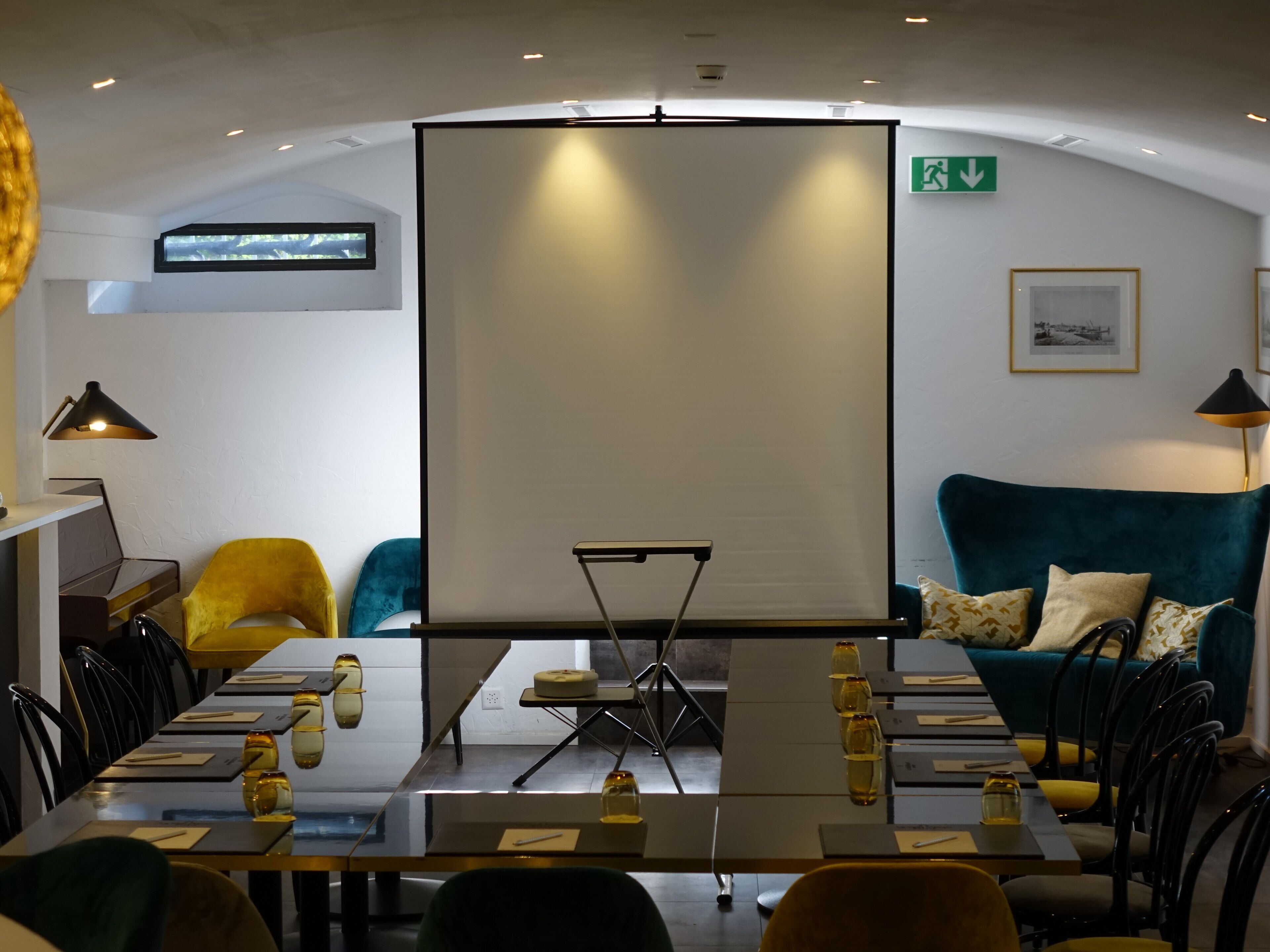 Un salón de conferencias con una pantalla grande, un piano, sillas amarillas y turquesas, y un sofá turquesa con cojines estampados.