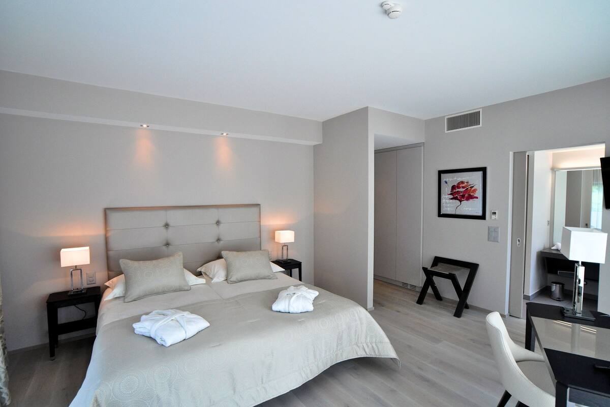 A modern hotel room with a large bed, two bedside tables with lamps, a desk with a chair, and a framed picture on the wall.