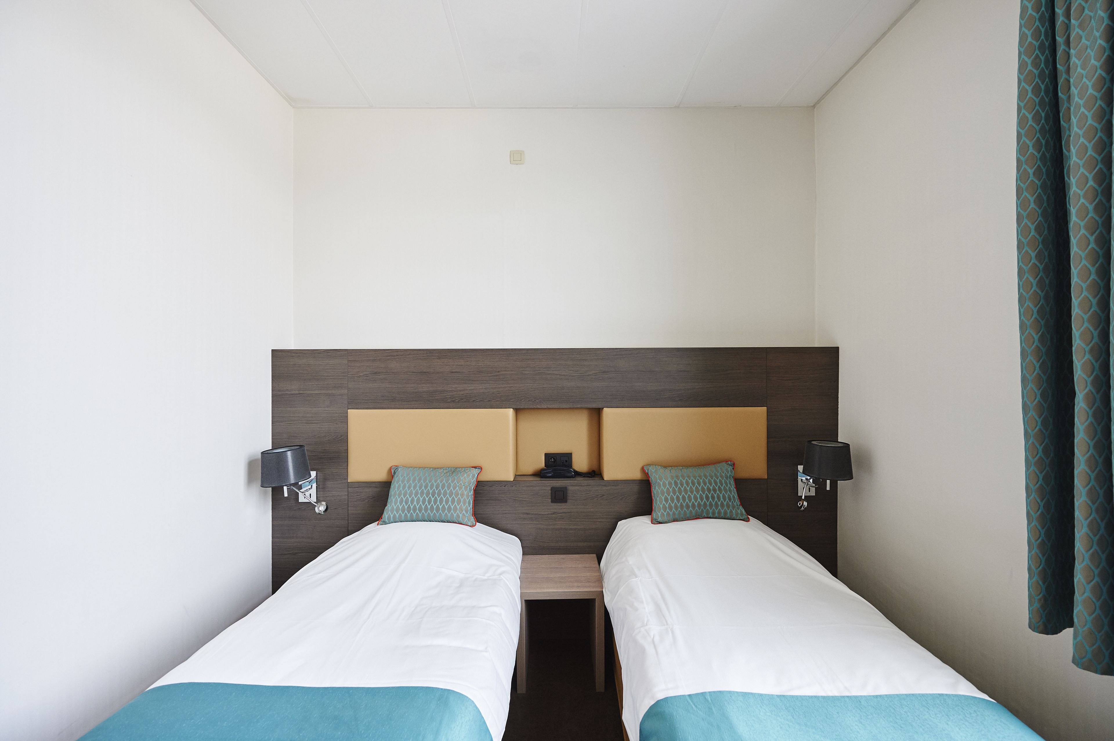 Habitación de hotel con dos camas individuales, cabecero de madera, lámparas de noche y una mesita entre las camas.