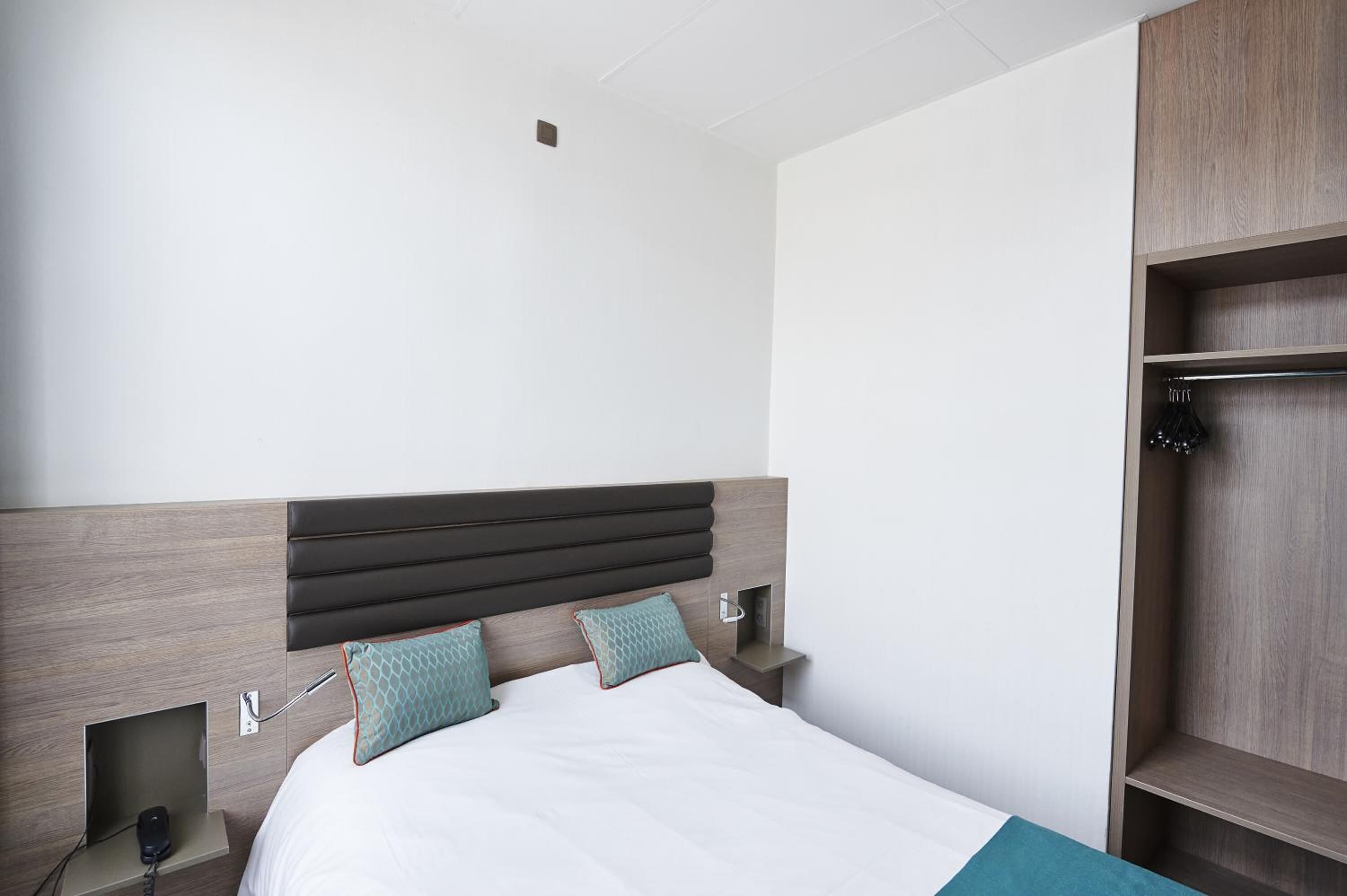 Une chambre d’hôtel moderne avec une tête de lit en bois, un lit recouvert de draps blancs, deux coussins turquoise, une table de chevet avec une lampe et une armoire intégrée.