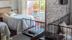 Une chambre d’hôtel avec un mur en pierre, un lit recouvert d’une courtepointe blanche, une table de chevet avec une télécommande, un plateau avec des tasses et une théière, et une fenêtre donnant sur l’extérieur.