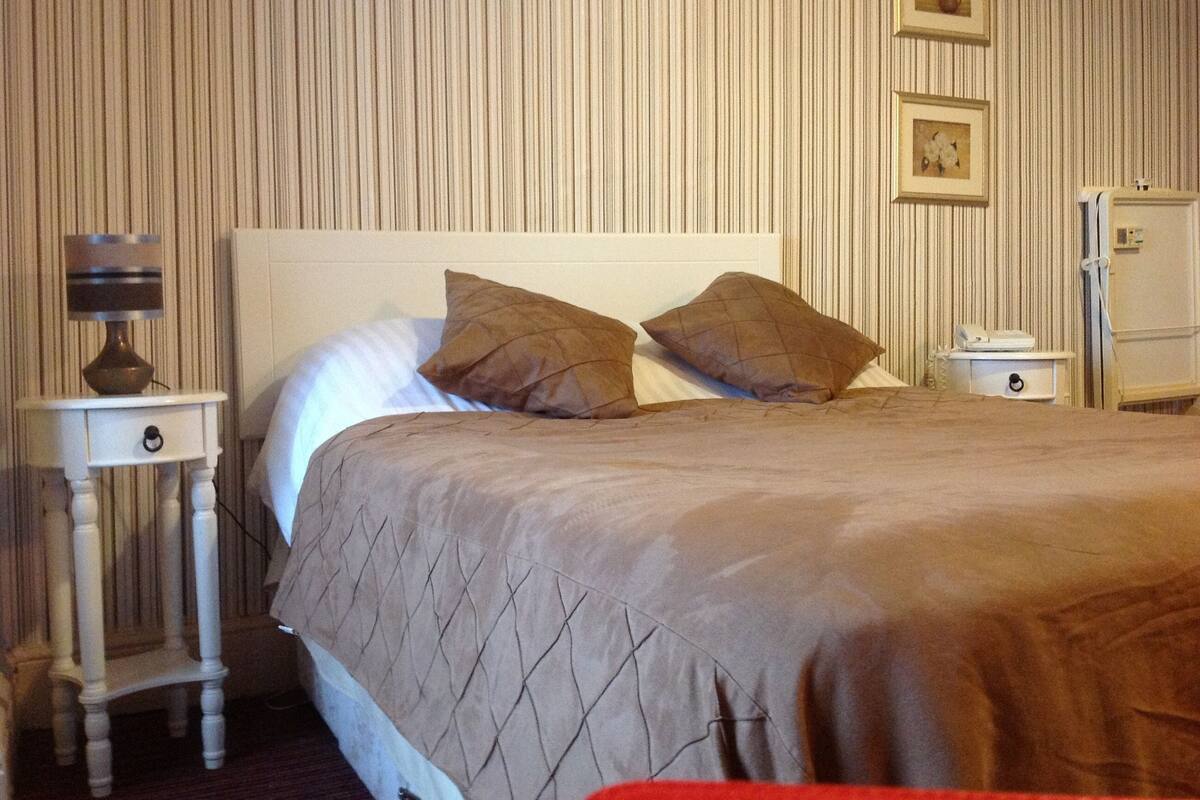 A neatly made bed with a quilted cover and two pillows, a bedside table with a lamp, and a nightstand with a phone.