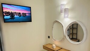 Un baño con una televisión mostrando una escena de atardecer, un tocador de madera con espejo, y una pequeña estantería con una vela.