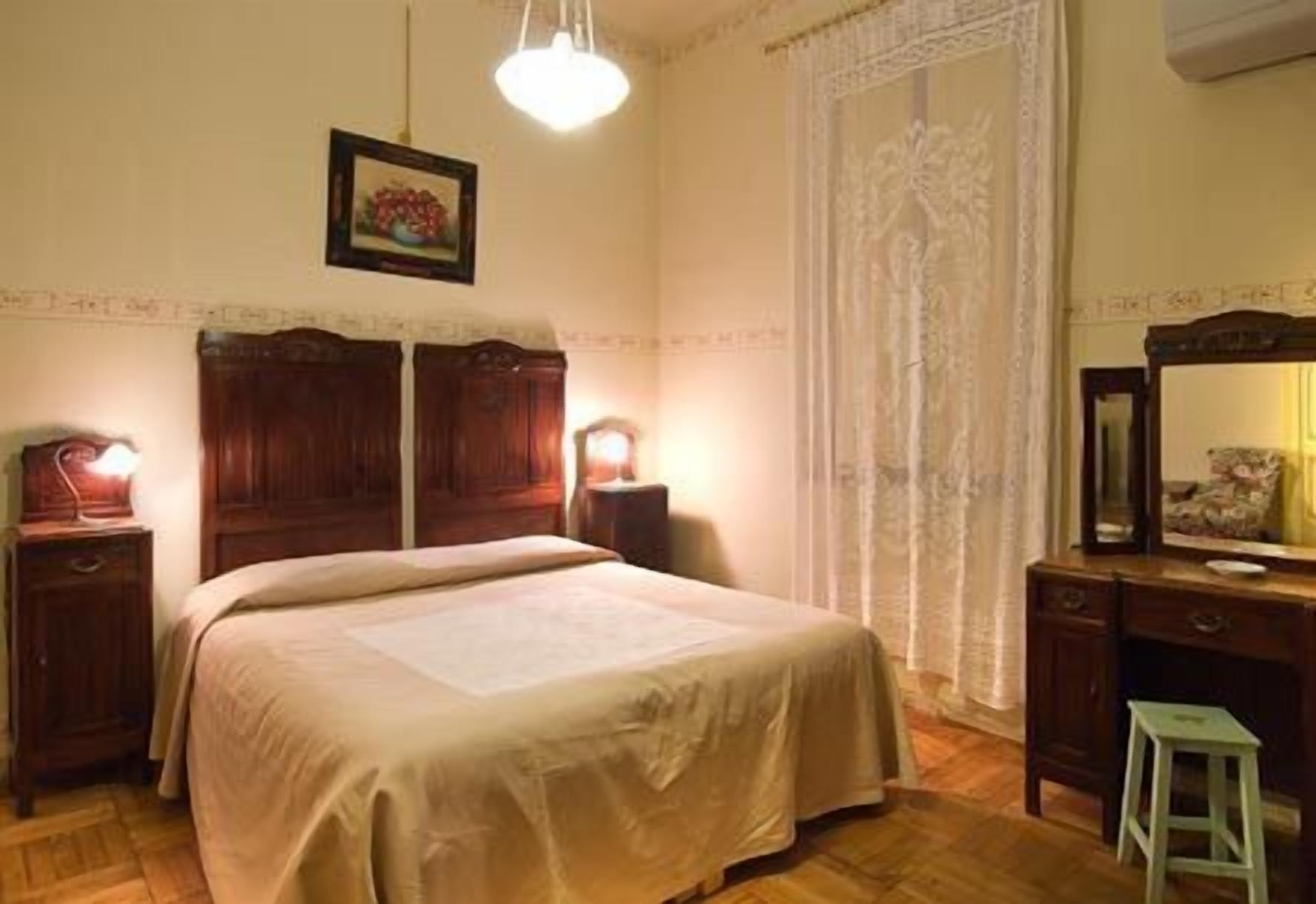 Un dormitorio con una cama grande, cabecera de madera, dos mesitas de noche con lámparas, un aparador, un espejo y un taburete pequeño.
