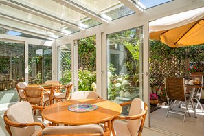 A sunlit patio with a round wooden table and wicker chairs, surrounded by glass doors leading to a lush garden. - Hotel Yacht Club (Marciana Marina)