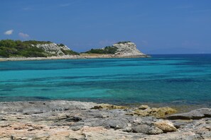 Een rotsachtige kustlijn met helder turquoise water en een kleine heuvel met schaarse begroeiing.