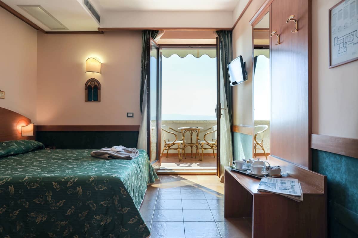 A hotel room with a bed, a desk with a chair, a television, and a balcony with a table and chairs.