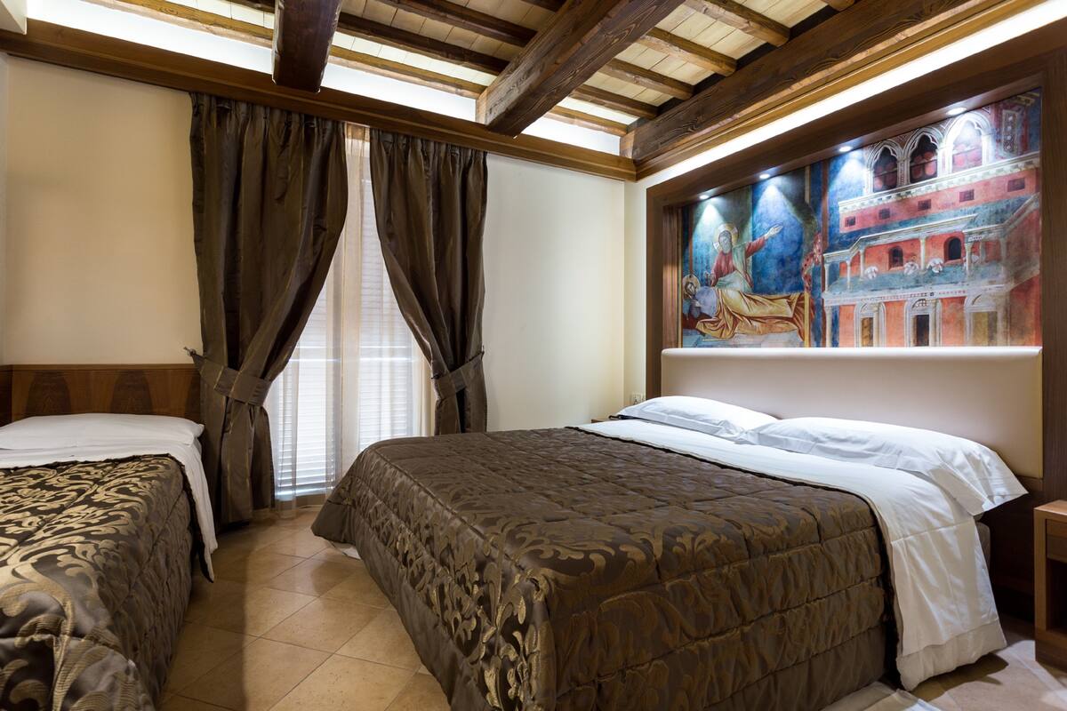 A hotel room with two beds, wooden beams on the ceiling, a large mural on the wall, and a window with curtains.