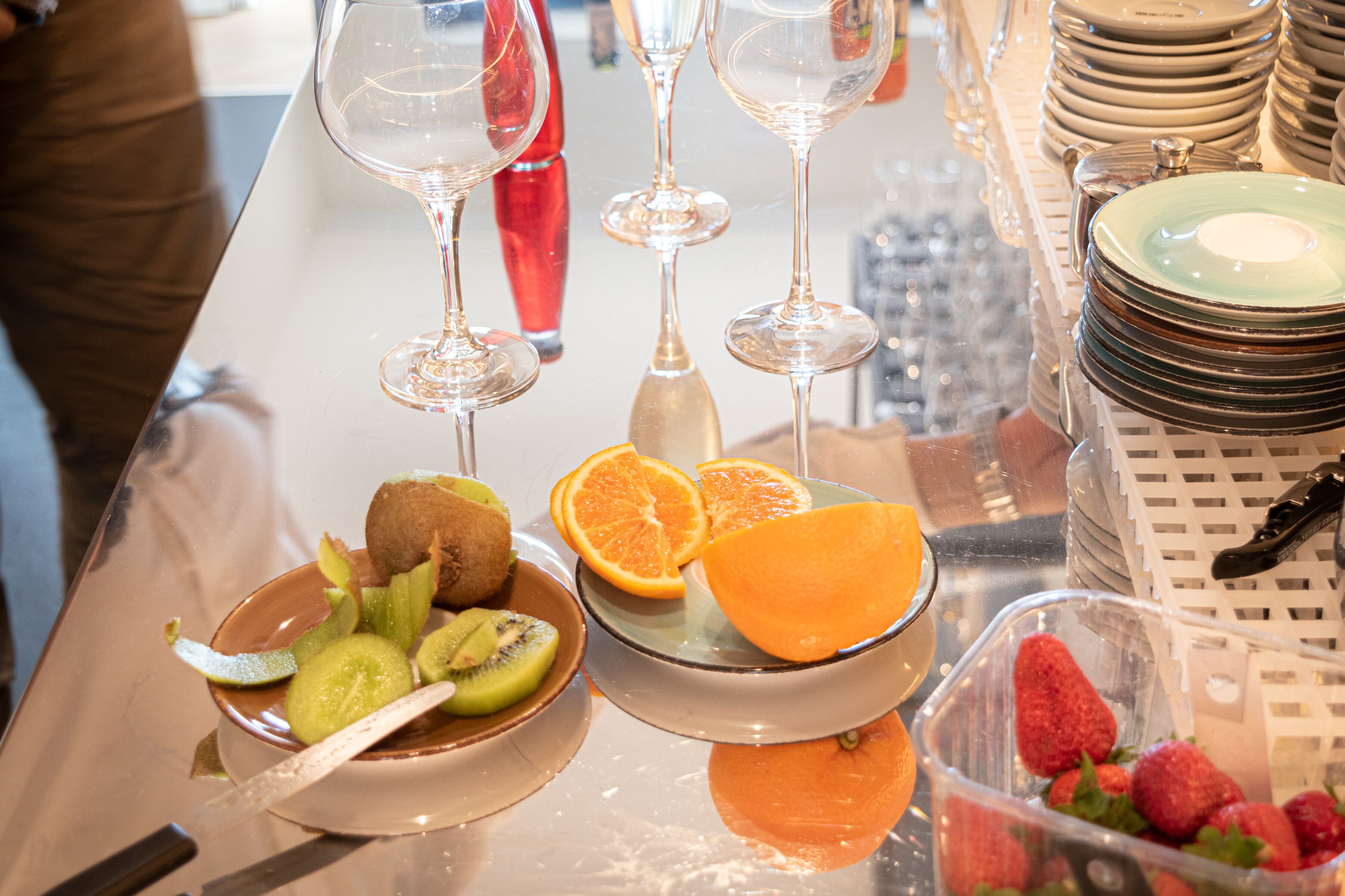 A table with various fruits, a bowl of kiwi, a glass of orange juice, and wine glasses.