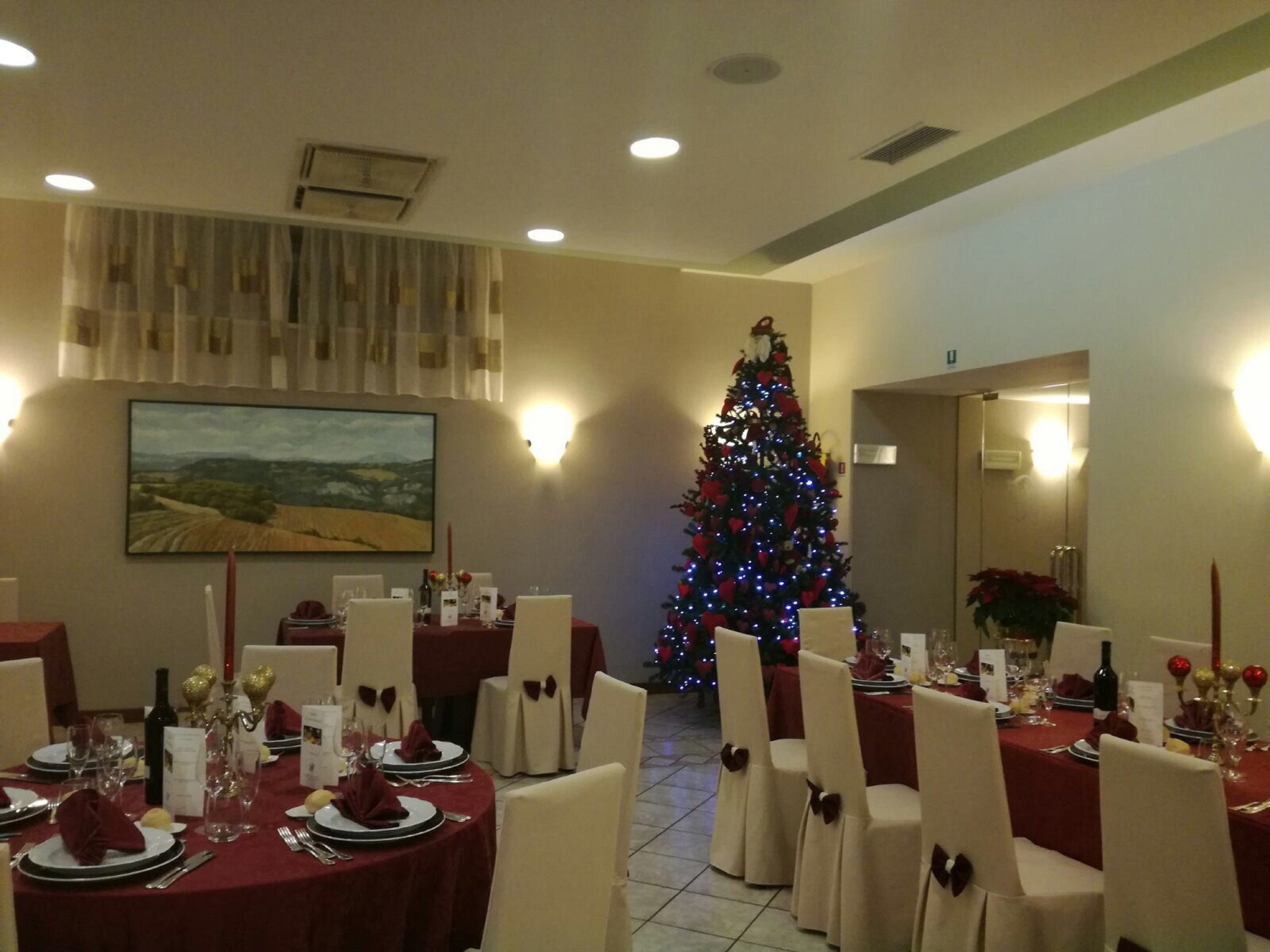 Un espace repas avec des tables rondes dressées pour le dîner, un sapin de Noël en arrière-plan et un tableau accroché au mur.