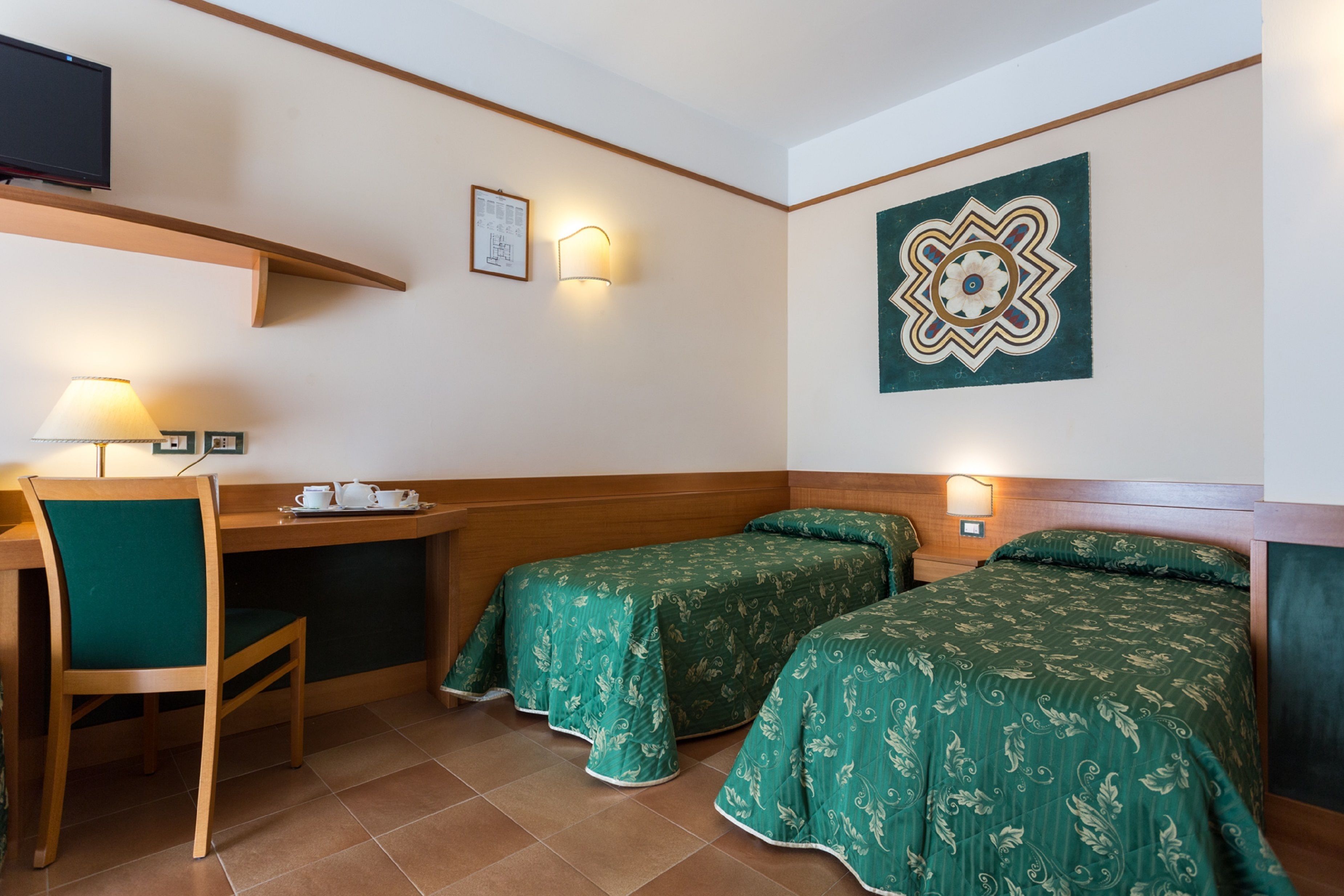 A hotel room with two single beds, a desk with a chair, a TV mounted on the wall, and a decorative wall piece.