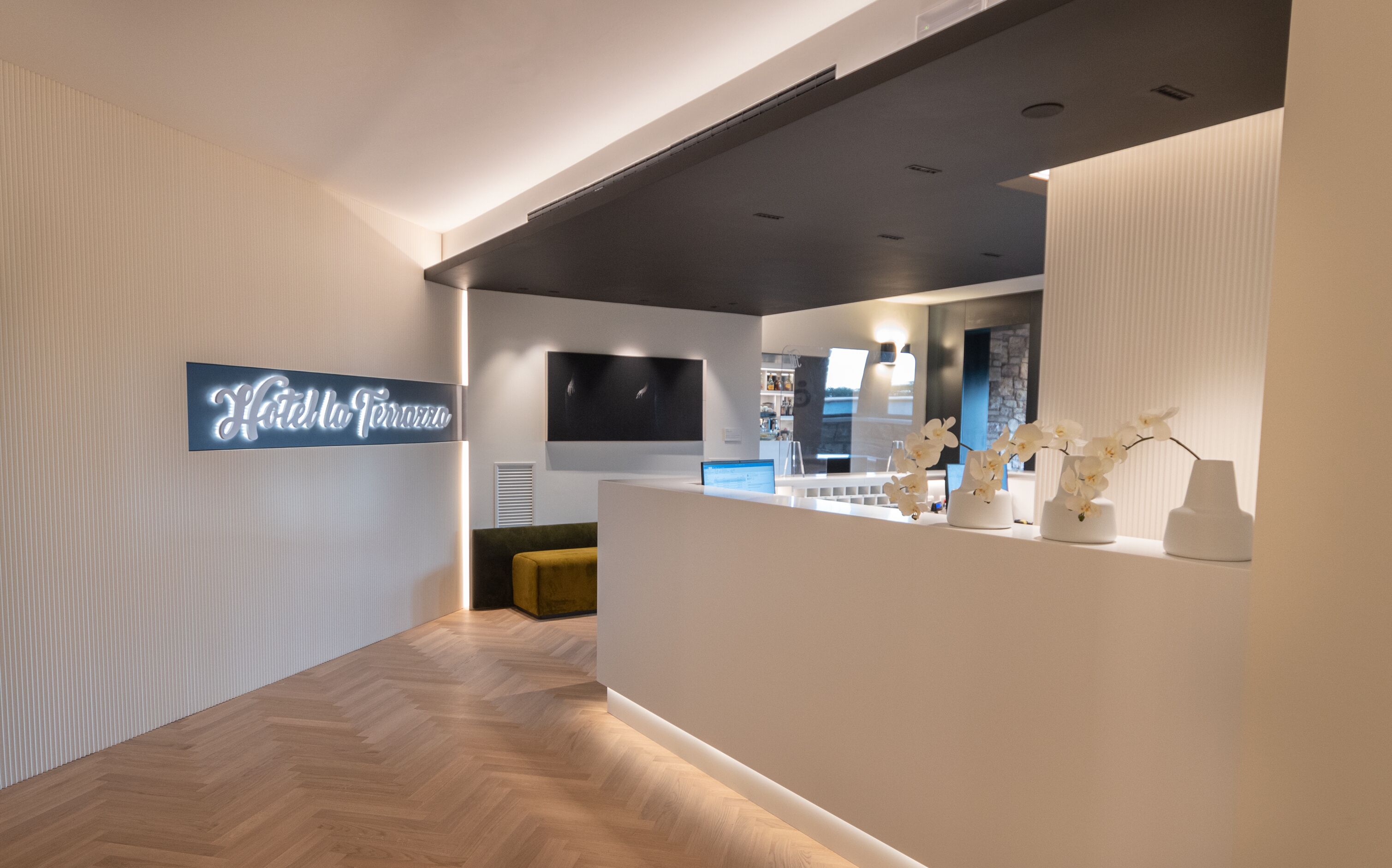 En modern hotellobby med ett fiskbensmönstrat trägolv, en receptionsdisk, en platt-TV och en skylt med texten ”Hotel Terrazzo”.