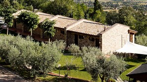 Edificio de piedra con tejado de tejas, rodeado de vegetación y olivos, con vistas a un paisaje lejano.
