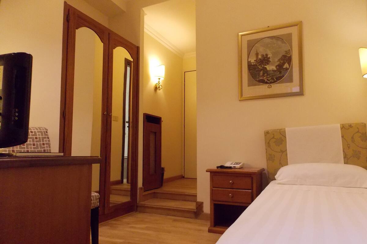 Hotellihuone, jossa on sänky, yöpöytä, lamppu, seinällä oleva taulu ja puinen ovi, joka johtaa toiseen huoneeseen.