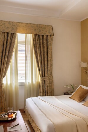 A hotel room with a neatly made bed, a bedside table with a glass of amber liquid, and a vase with a single white flower. - Hotel Cortina (Rome)