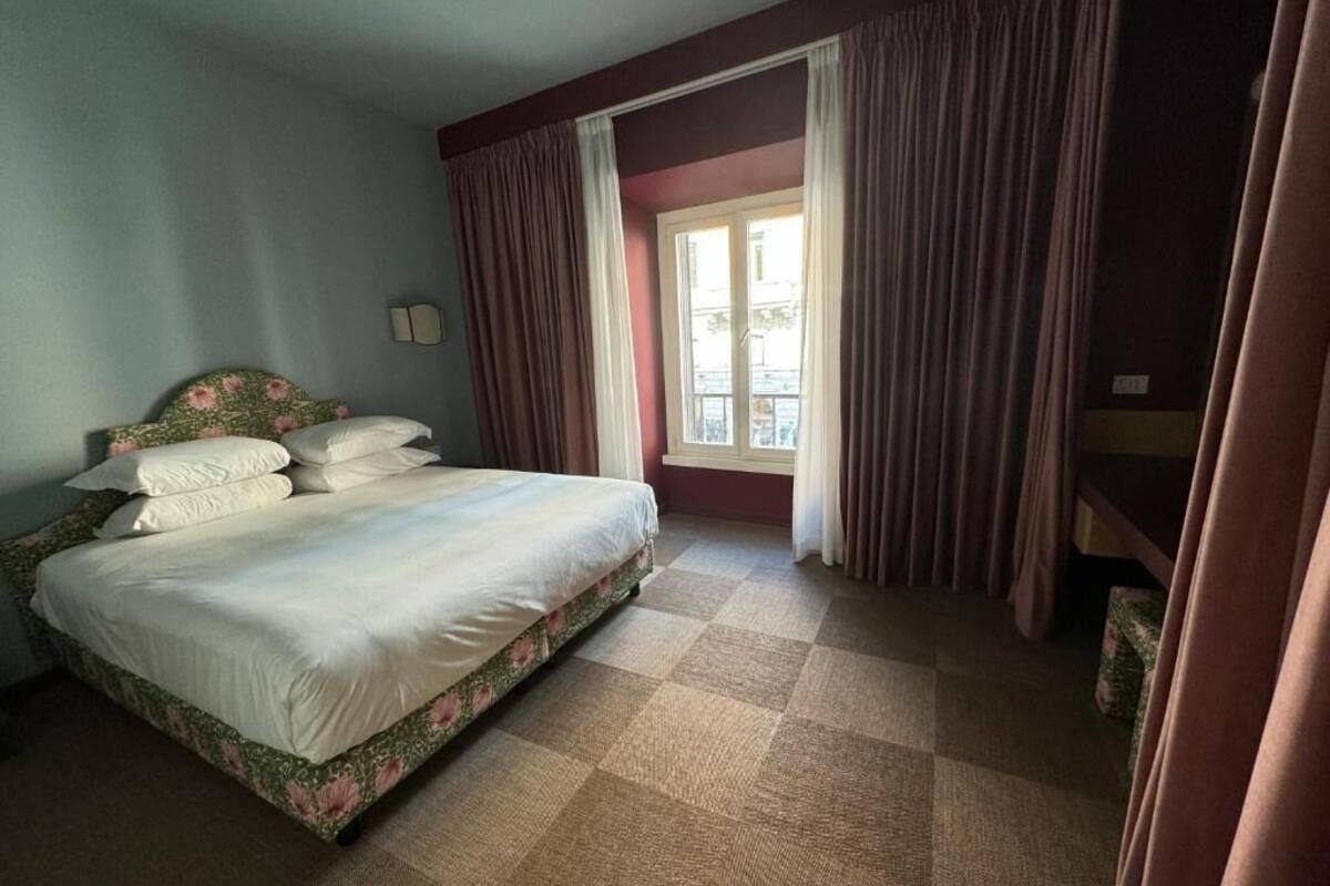 Hotellihuone, jossa on sänky, verhot, ikkuna ja ruutukuviollinen lattia.
