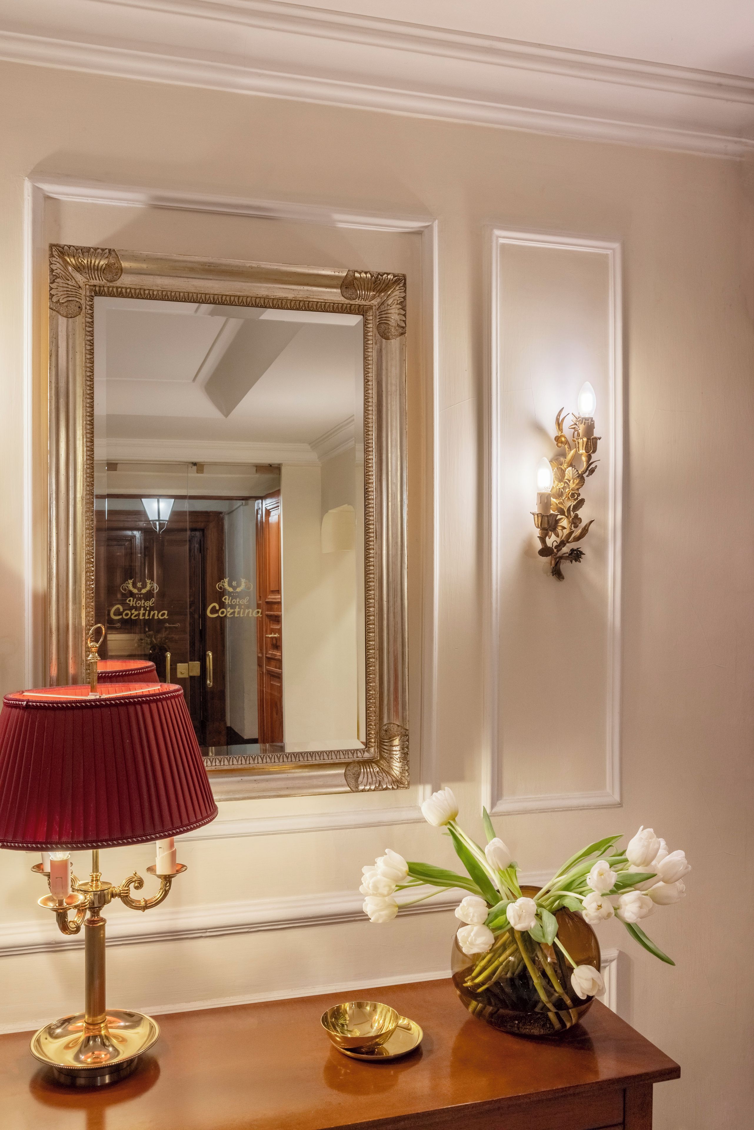 Une pièce avec un grand miroir décoratif, une lampe rouge, un chandelier doré et un vase rempli de fleurs blanches, le tout posé sur une table en bois.