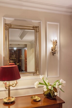 A room with a large ornate mirror, a red lamp, a gold candelabra, and a vase with white flowers on a wooden table. - Hotel Cortina (Rome)