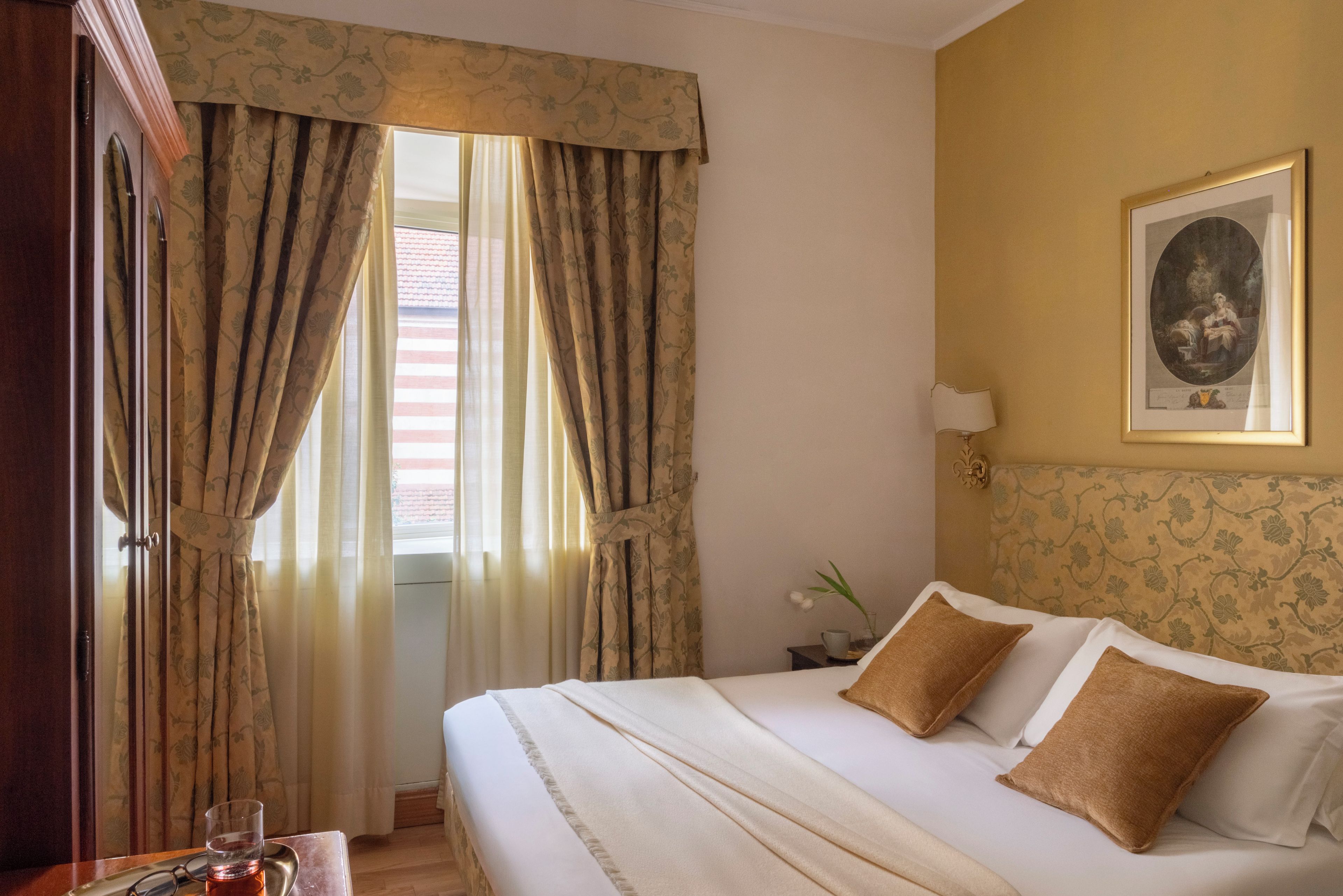 Une chambre d’hôtel avec un lit, des rideaux, un tableau encadré au mur et une petite table sur laquelle se trouve une tasse.