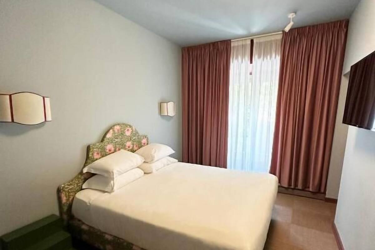 Hotellihuone, jossa on sänky, televisio, valaisin ja ikkuna, jossa on verhot.