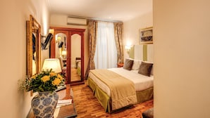 Une chambre d’hôtel avec un plancher en bois, un lit recouvert d’une couvre-lit beige et verte, une table de chevet avec une lampe, un miroir et un vase avec des fleurs jaunes.