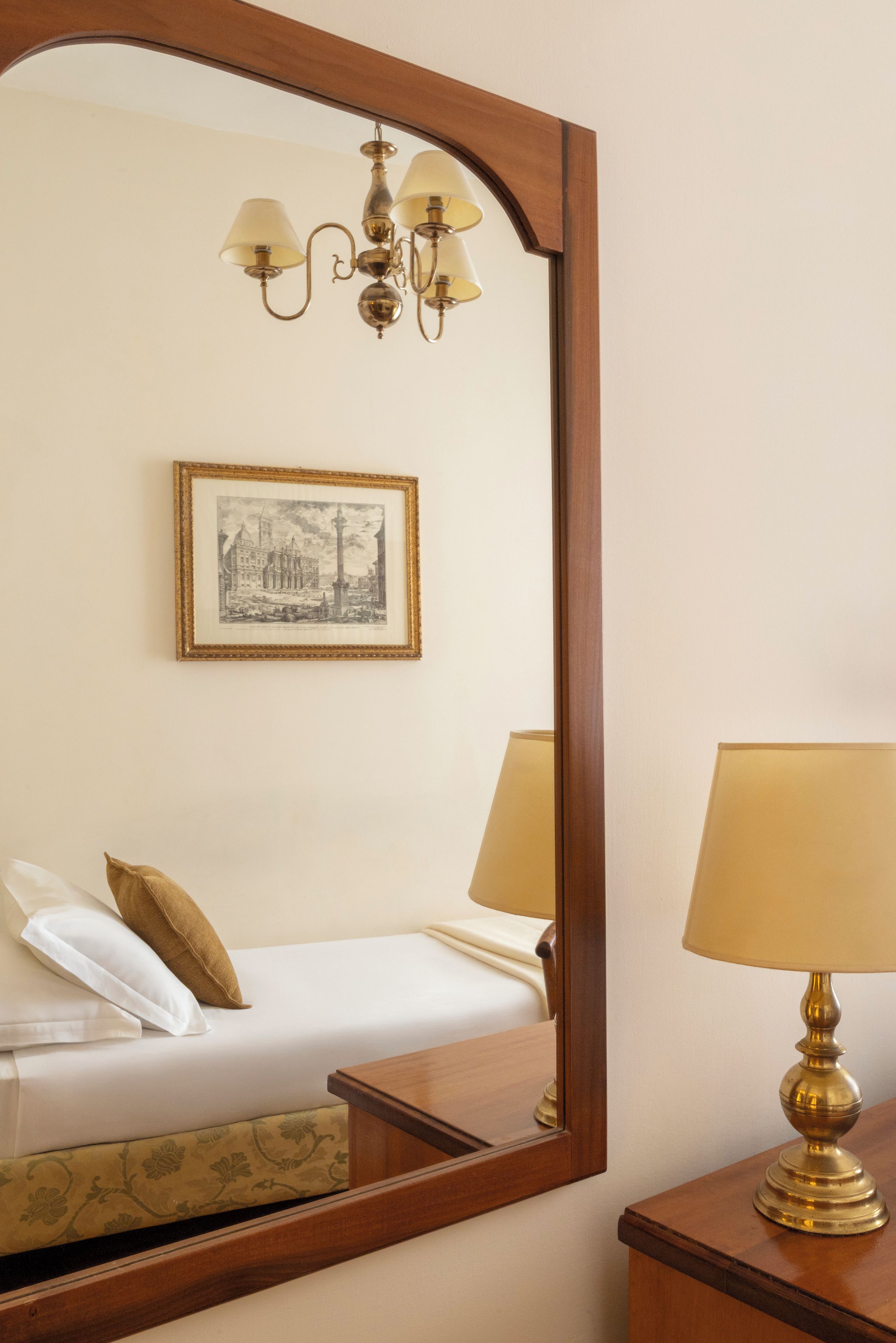 Quarto de hotel com cama, mesa de cabeceira, abajur, quadro pendurado na parede e espelho.