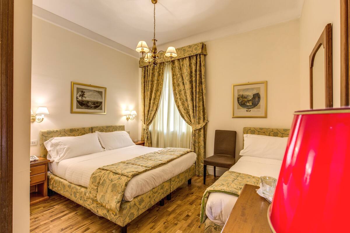 Hotellihuone, jossa on kaksi sänkyä, puulattia, kattokruunu ja seinillä olevat taulut.