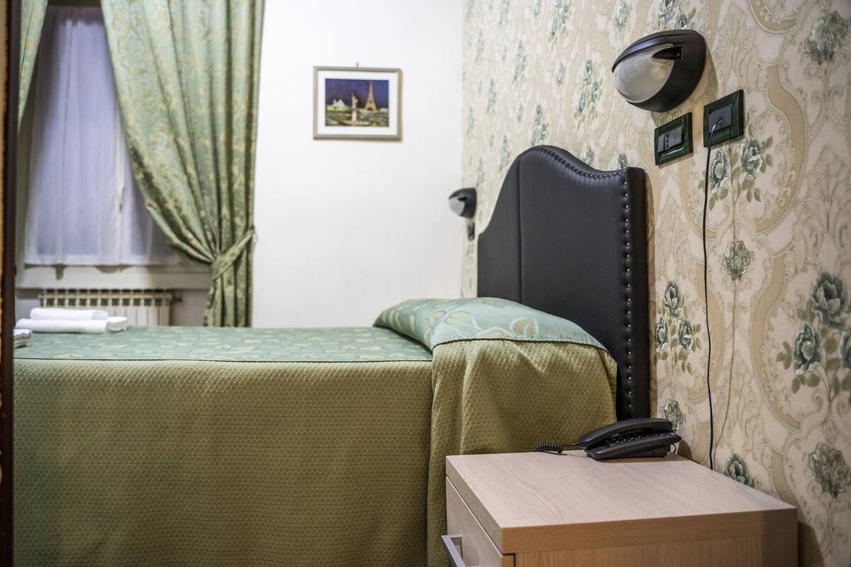 A room with a bed, a bedside table with a telephone, a wall-mounted lamp, and a picture on the wall.