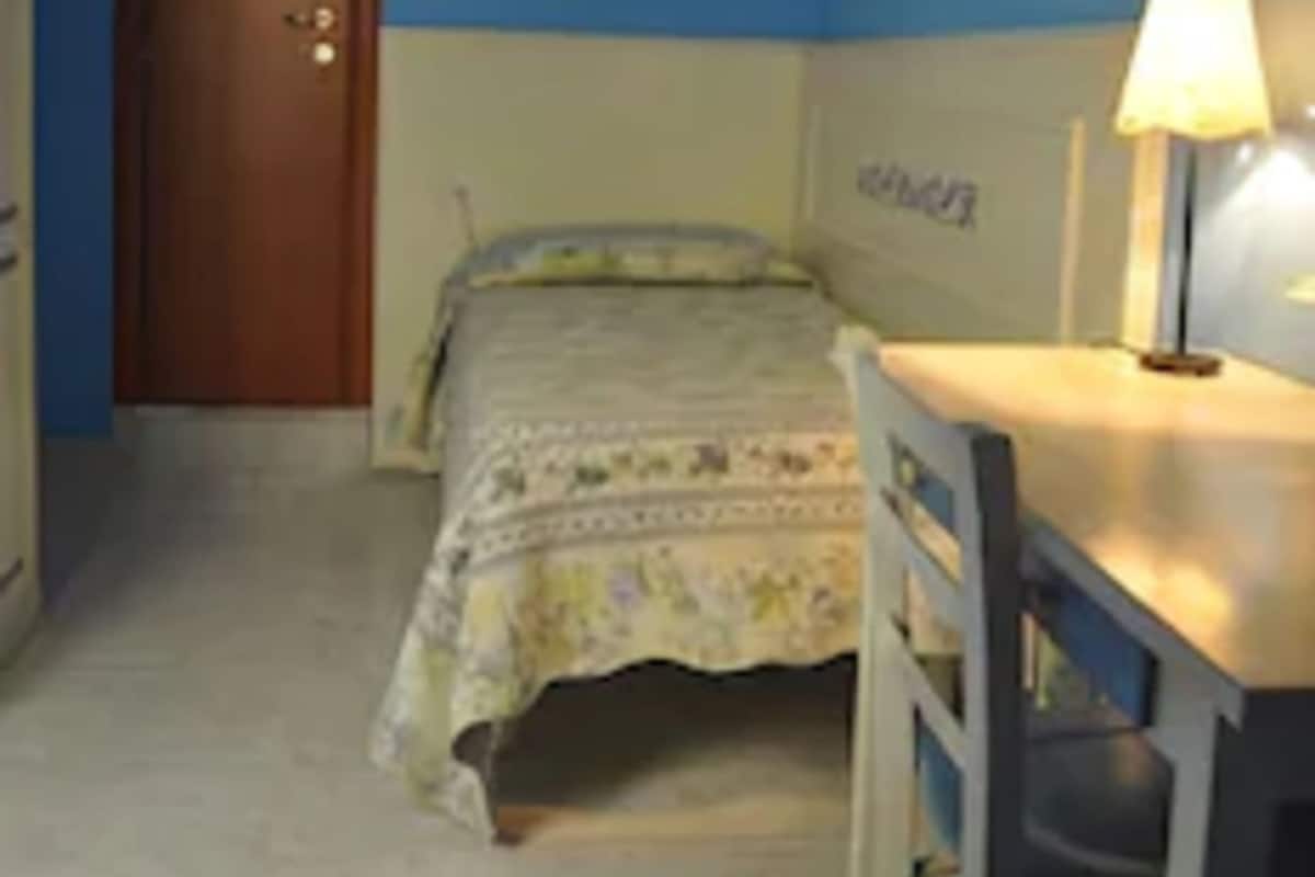 Un dormitorio con cama, escritorio, silla, lámpara y puerta.