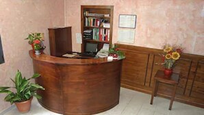 Una reception con una scrivania, un computer, un telefono e una piccola libreria. Sono presenti piante in vaso e una panca di legno con un vaso di fiori.