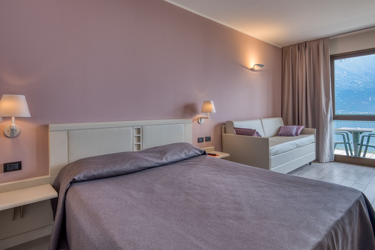 A hotel room with a bed, a sofa, two bedside tables with lamps, a window with curtains, and a view of the sea.