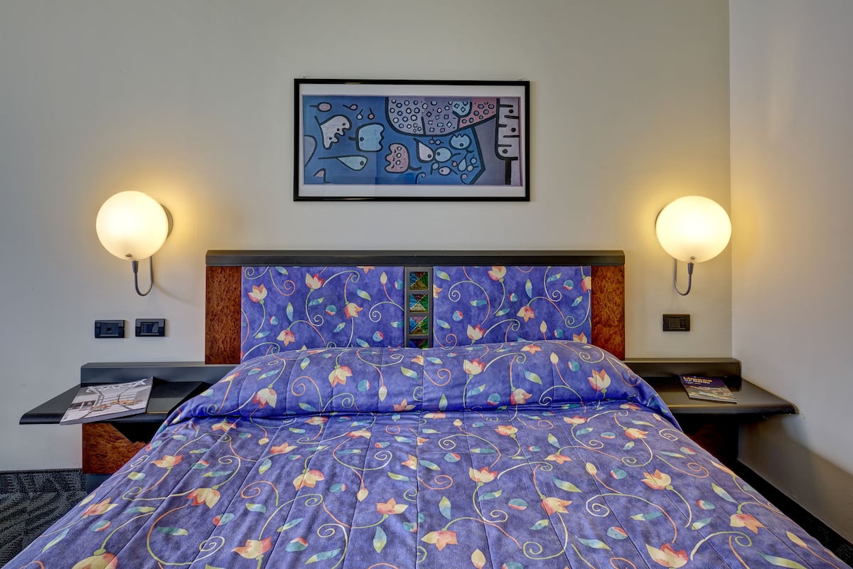 A hotel room with a bed, two bedside tables, a framed artwork above the bed, and wall-mounted reading lights.